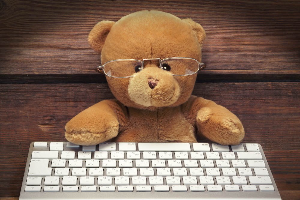 Cute Teddy Bear With A Wireless Keyboard In Front Of An Imaginary Computer Screen At Wooden Wall In Lanters Light. Internet Surfing Or Computer Work Concept