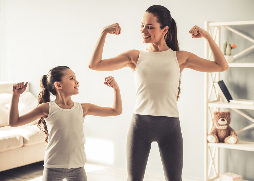 604202222 Beautiful young woman and charming little daughter are showing their biceps and smiling while working out at home