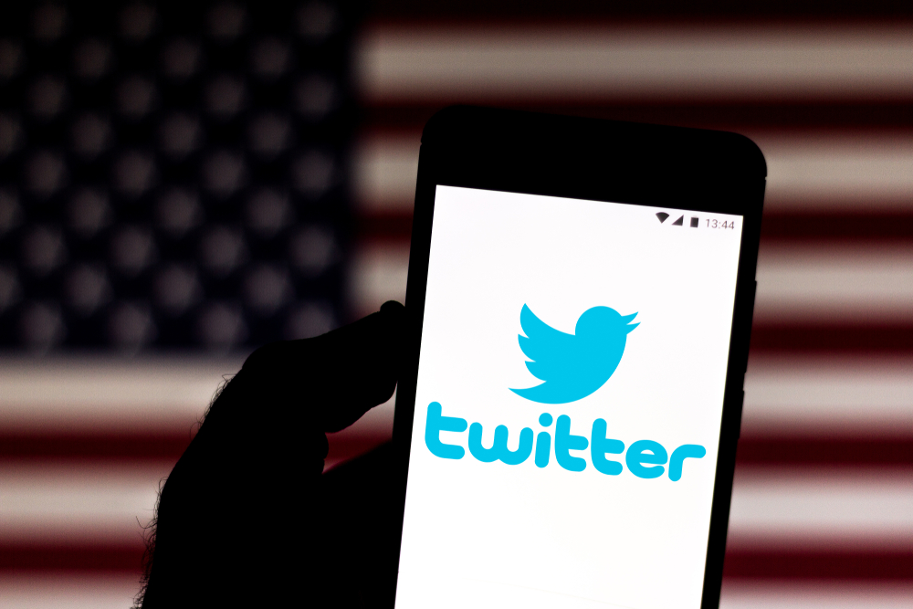 Shutterstock_ 1449809192 Twitter logo is displayed on a smartphone. In the background, flag of the United States of America.