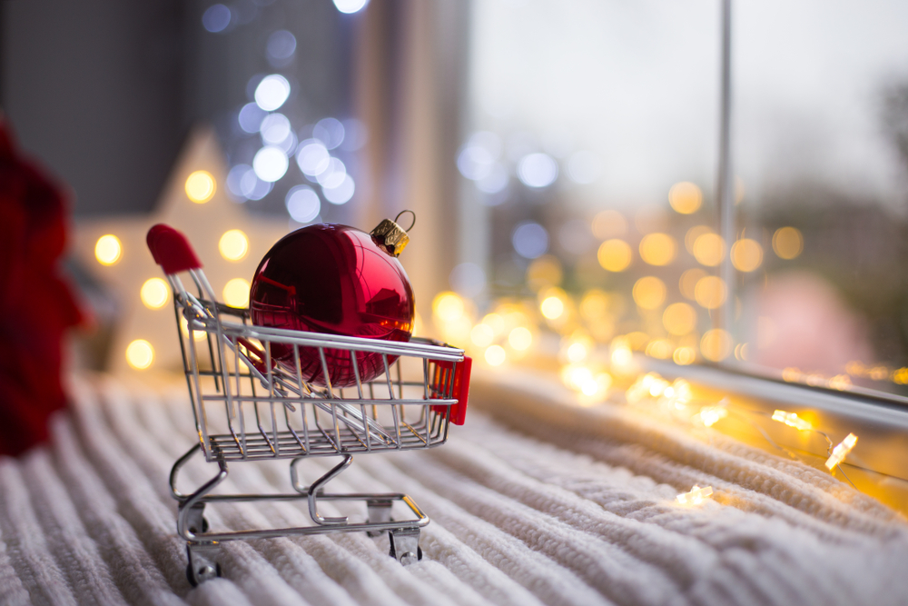 Shutterstock_1185684814 Red Christmas ball in toy supermarket trolley in daylight with warm garland bokeh on background. Festive magic lights. New Year shopping concept.