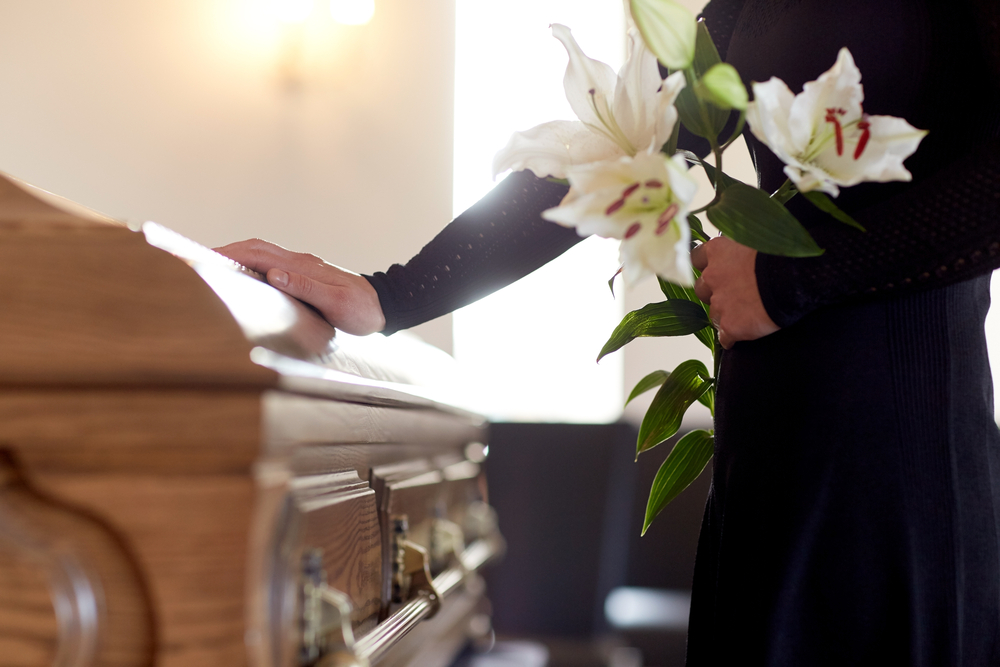 Shutterstock_664197991 people and mourning concept - woman with white lily flowers and coffin at funeral in church