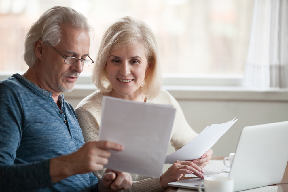 Shutterstock_1185179278 Happy old middle aged couple holding reading good news in document, smiling senior mature family excited by mail letter, checking paying domestic bills online on laptop, discussing budget planning