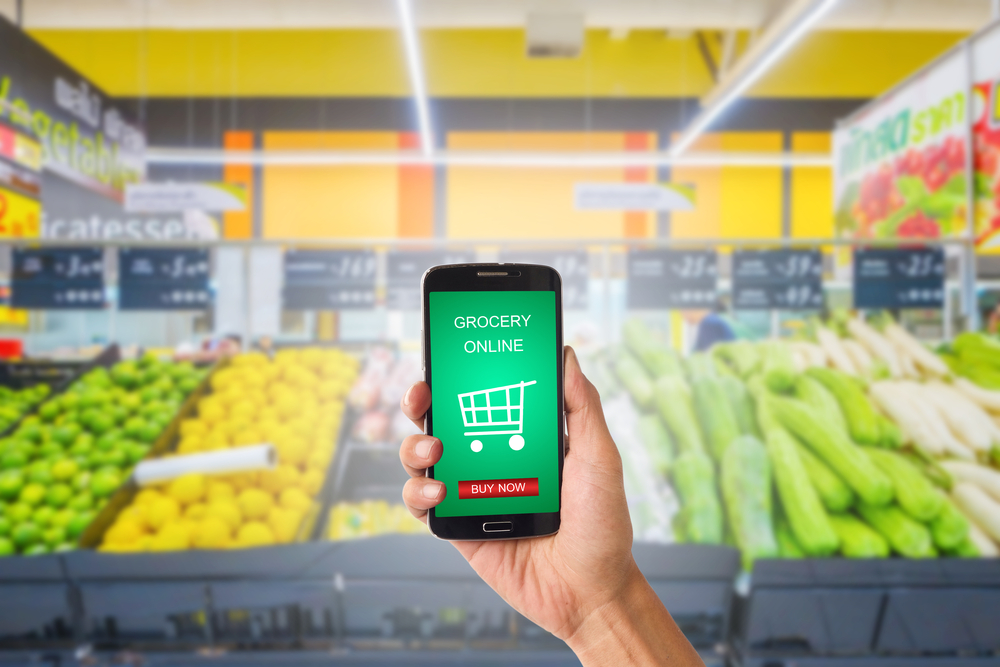 Shutterstock_555054529 Hand holding mobile with grocery online on screen with blur supermarket background