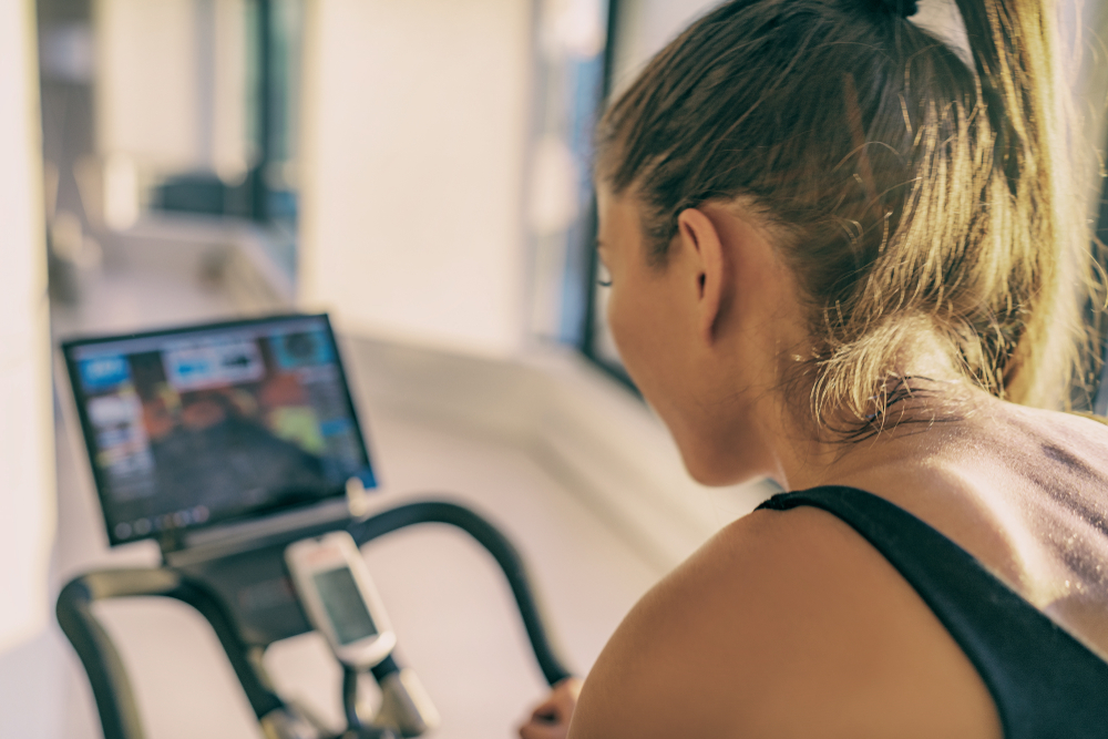 Shutterstock_1297790632 Smart fitness home workout biking screen with online classes woman training on stationary bike equipment indoors for biking exercise. Indoor cycling. Focus on the sweat on person's back.