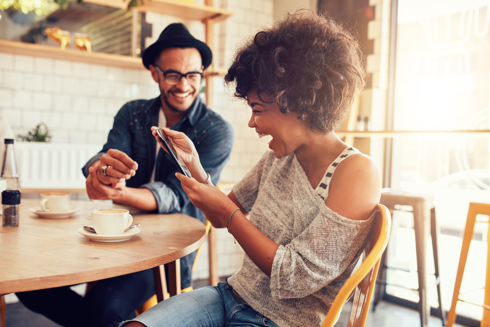 Shutterstock_380240200 Portrait of smiling young woman at a cafe table looking at digital tablet with a friend sitting by.