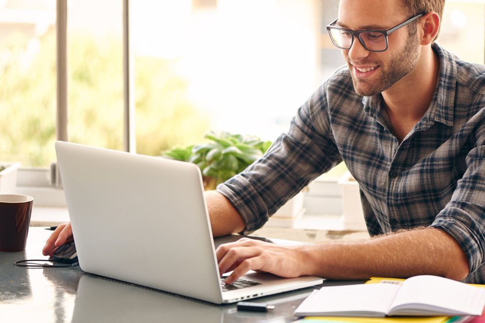 Shutterstock_272163653 Happy young man, wearing glasses and smiling, as he works on his laptop to get all his business done early in the morning with his cup of coffee