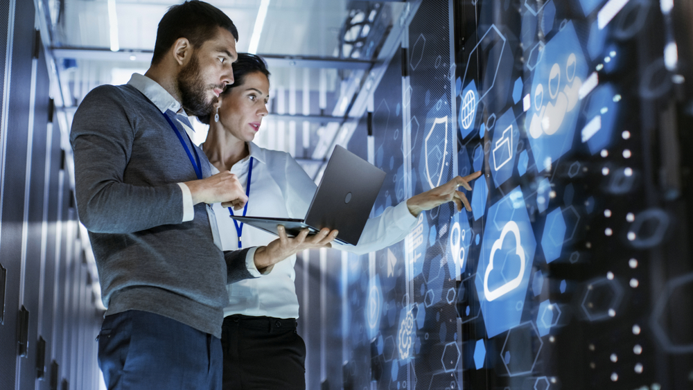 Shutterstock_1062915392  Male IT Specialist Holds Laptop and Discusses Work with Female Server Technician. They're Standing in Data Center, Rack Server Cabinet with Cloud Server Icon and Visualization.