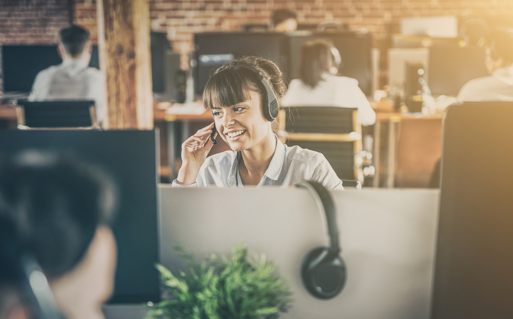 Shutterstock_739359772  Call center worker accompanied by her team. Smiling customer support operator at work. Young employee working with a headset.