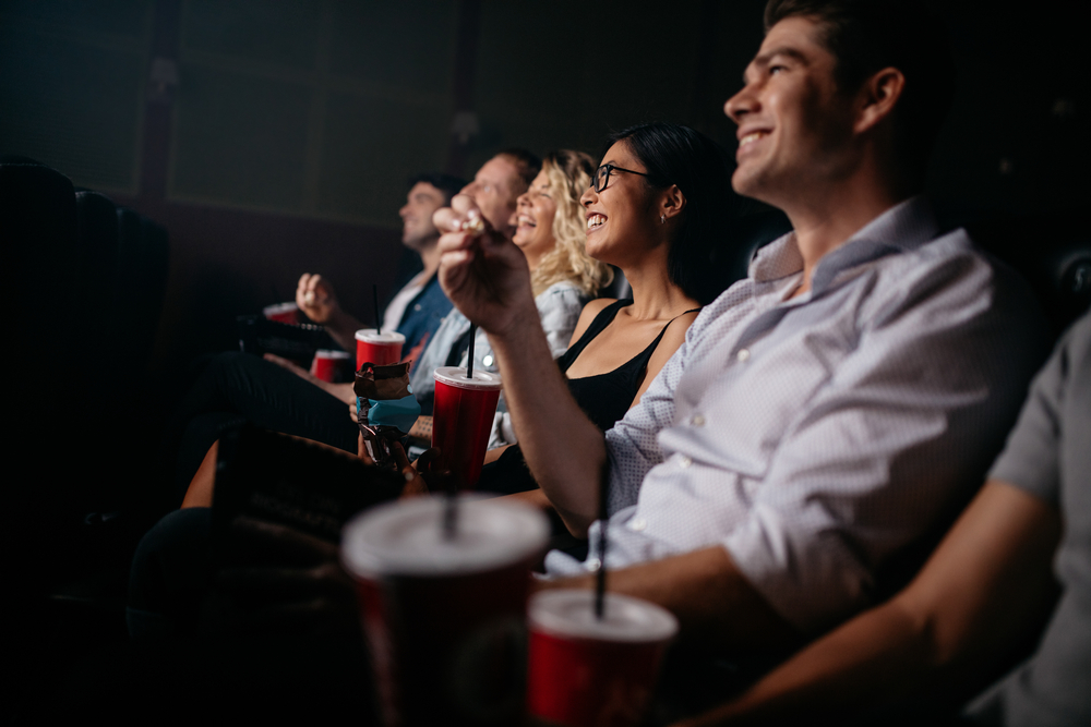 Shutterstock_518102128 Group of people in theater with popcorn and drinks. Young friends watching movie in cinema.