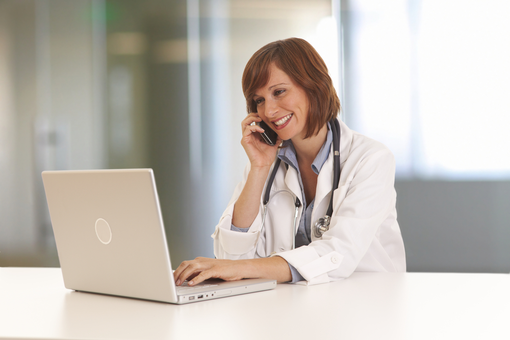 Shutterstock_71805475  Portrait of young woman doctor in white coat at computer using phone