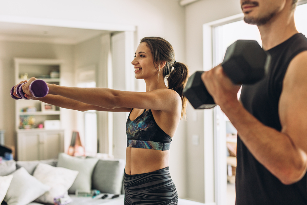 Shutterstock_1445395694 Healthy young couple doing exercises with dumbbells at home. Fit young woman with man doing weights workout indoors in living room.