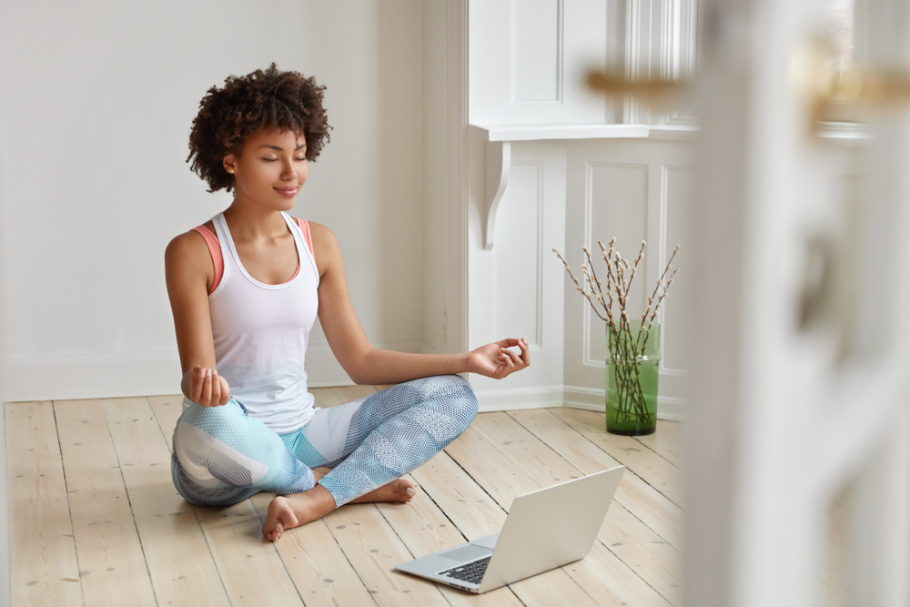 Shutterstock_1226534068 Calm lady with Afro hairstyle, dressed in sportswear, meditates on floor in empty room, listens spiritual practices lessons on laptop computer, poses in lotus pose, tries to relax. Yoga concept