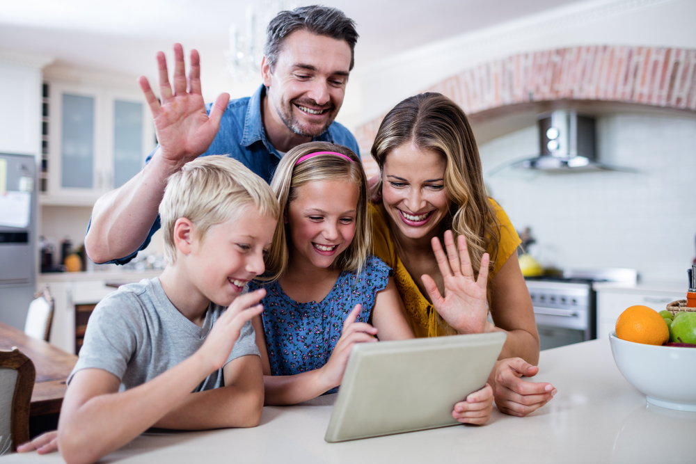 Shutterstock_572378356 Parents and kids waving hands while using digital tablet for video chat in kitchen