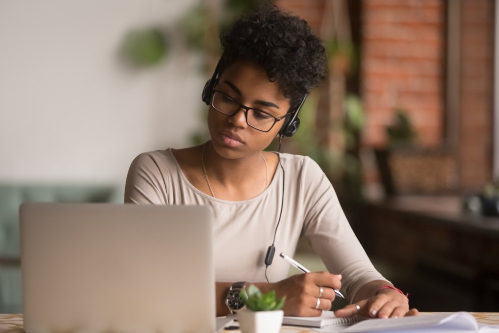 Shutterstock_1375976735 Focused mixed race woman wearing headphones watching webinar write notes study online with online teacher, african female student learning language computer course on laptop listen translate lecture