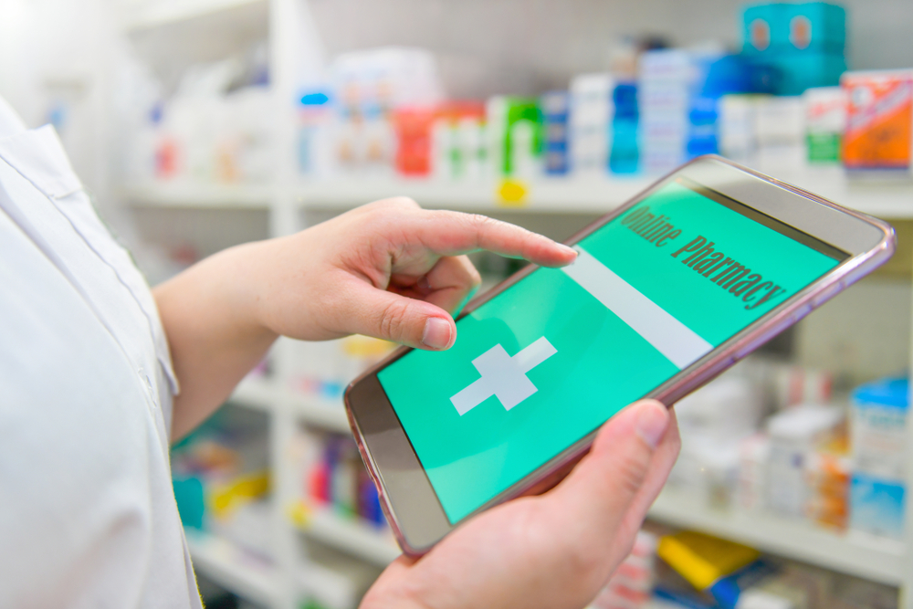 Shutterstock_1471304741 Pharmacist holding touchpad for search bar on display in pharmacy drugstore shelves background.Online medical concept.