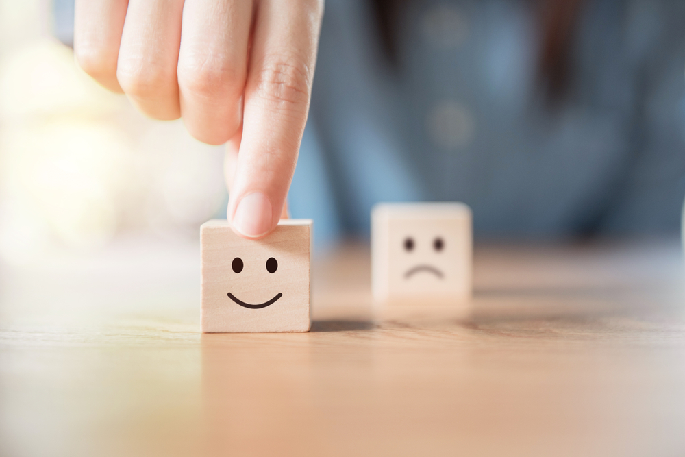 Shutterstock_1170951868 Close up customer hand choose smiley face and blurred sad face icon on wood cube, Service rating, satisfaction concept.