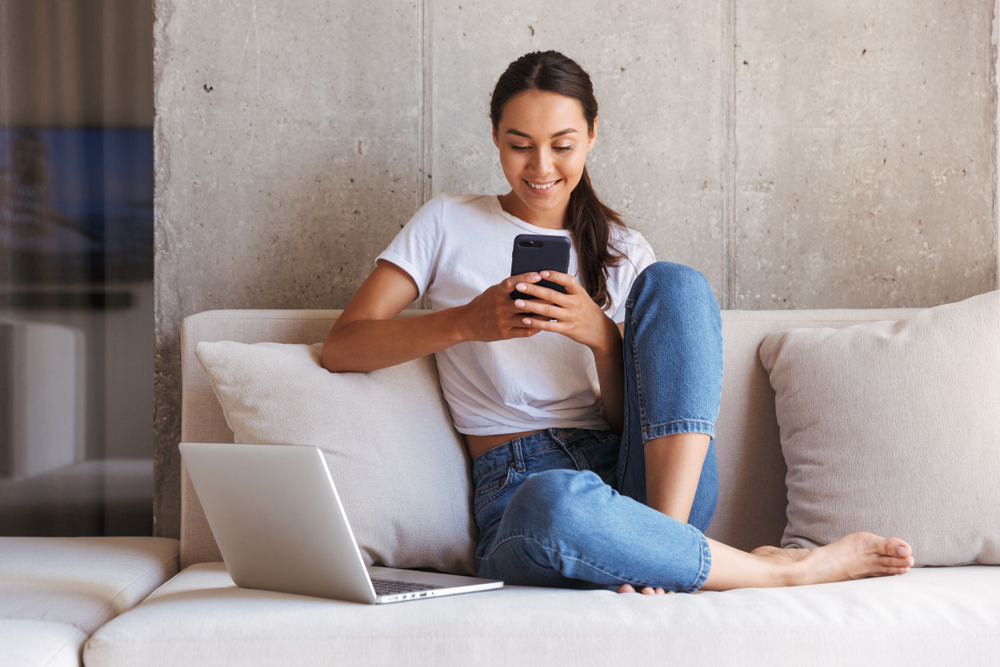 Shutterstock_1125823856 Happy young asian woman using mobile phone while sitting a couch at home with laptop computer