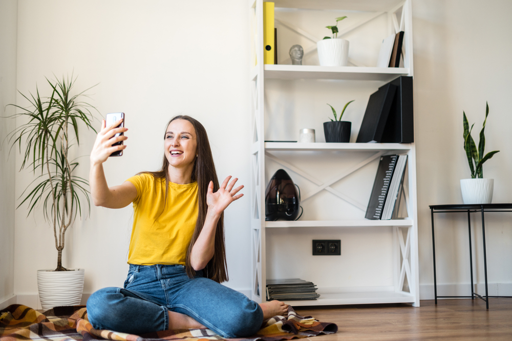 Shutterstock_1687992496 Phone video call. A young woman communicates via video, zoom. She is sitting on the floor in a Scandinavian style room.