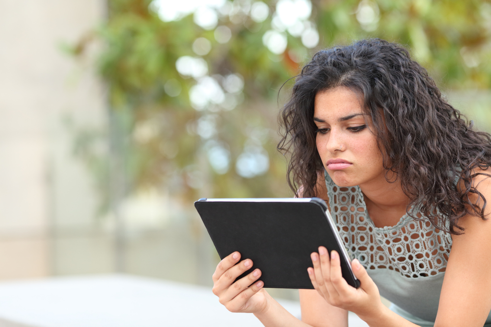 shutterstock_1276121887  Bored woman watching media content in a tablet sitting in a park