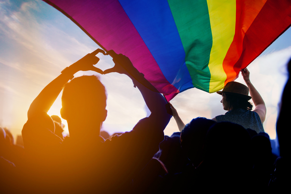 Shutterstock_1177679803 Pride community at a parade with hands raised and the LGBT flag.