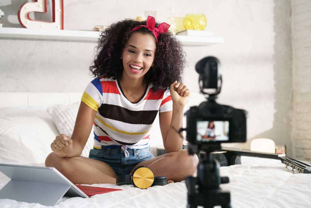 Shutterstock_1094656709 Happy girl at home speaking in front of camera for vlog. Young black woman working as blogger, recording video tutorial for Internet