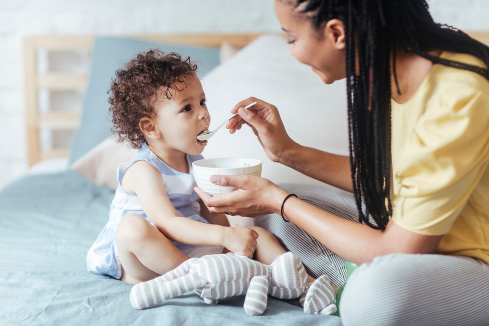 Shutterstock_770901967 African woman giving her baby to eat and sitting in bedroom.