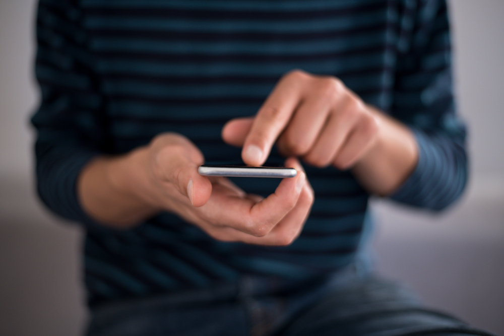 Shutterstock_1138238765 Young man using apps on a touchscreen smartphone - Hands close-up, focus on phone - Blue jeans and t-shirt - Concept for using technology, shopping online, using mobile apps, texting, phone addiction