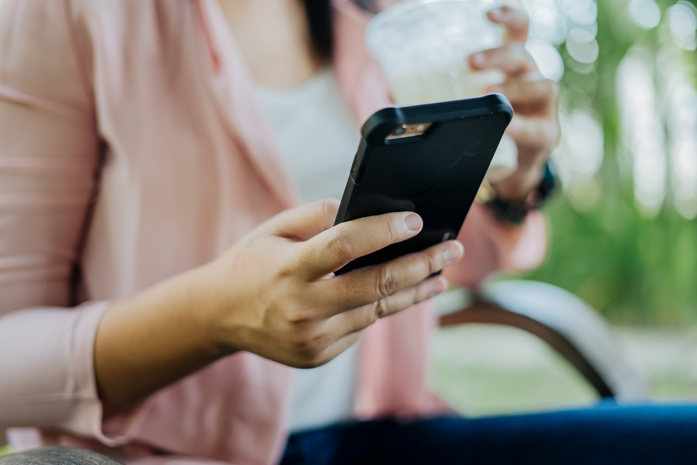 Shutterstock_541239523 Asian Woman typing text message on smart phone in a cafe. Cropped image of young woman sitting at a chair with a coffee using mobile phone.