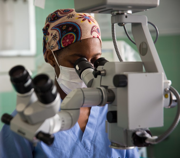 Shutterstock_1420207253 African Female doctor looking through Operating microscope equipment with face mask Cape Town South Africa.