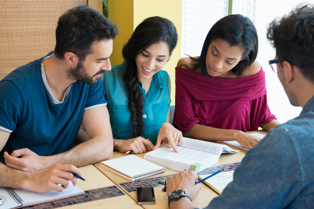 Shutterstock_313677374 Closeup shot of young man and woman discussing on note. Happy and smiling student studying at the library. Multi ethnic group studying on book for the next exam.