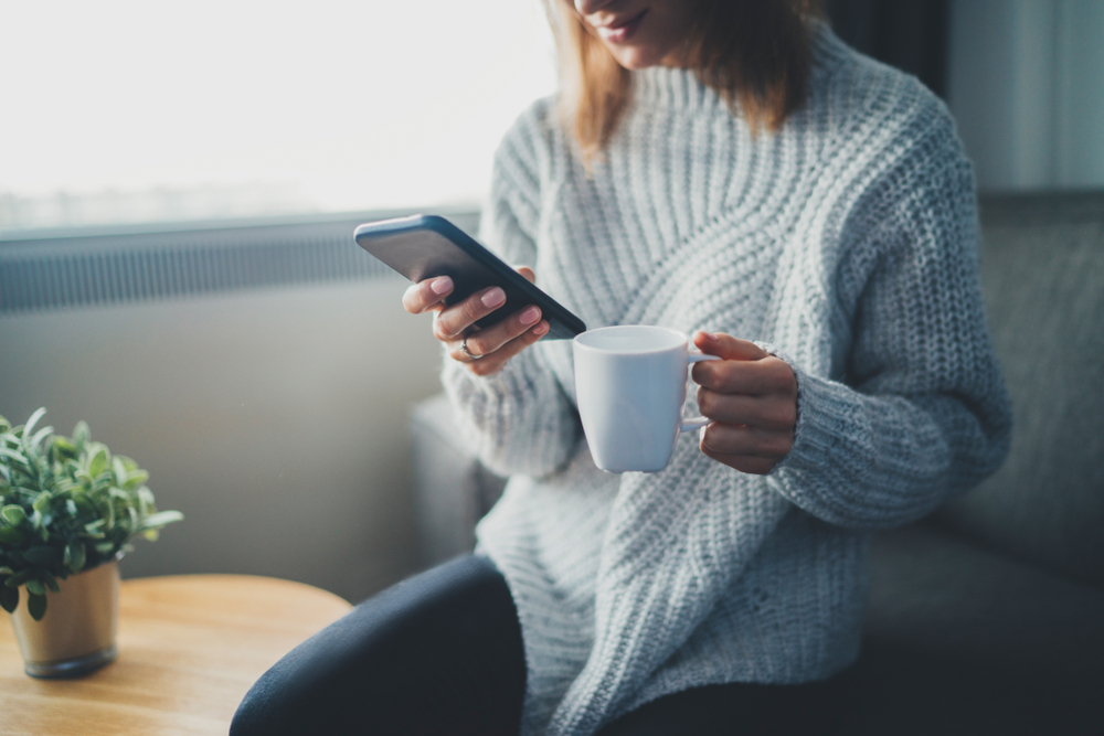 Shutterstock_794105284 Happy smiling hipster girl using smartphone device while chilling at home wearing cozy knitted sweater, attractive young woman chatting with friends at social network while sitting on sofa at home