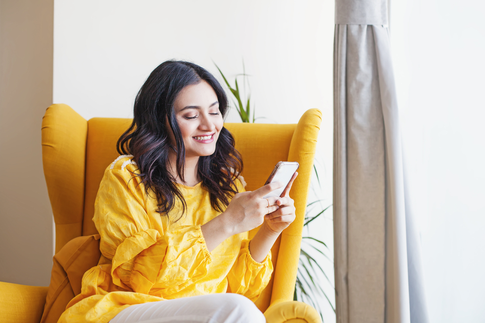 Shutterstock_1453316822 Young beautiful Indian woman using her mobile phone while sitting in the armchair at home