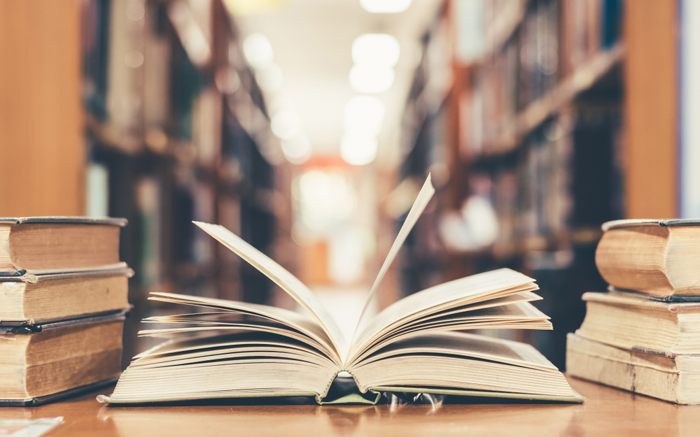 Shutterstock_794015686 Education learning concept with opening book or textbook in old library, stack piles of literature text academic archive on reading desk and aisle of bookshelves in school study class room background