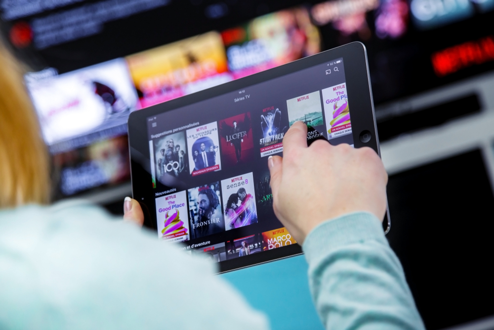 Shutterstock_1009681927 Benon, France - January 21, 2018: Woman Holding a touch pad and switching channels on France Netflix HomePage. with TV set on background.