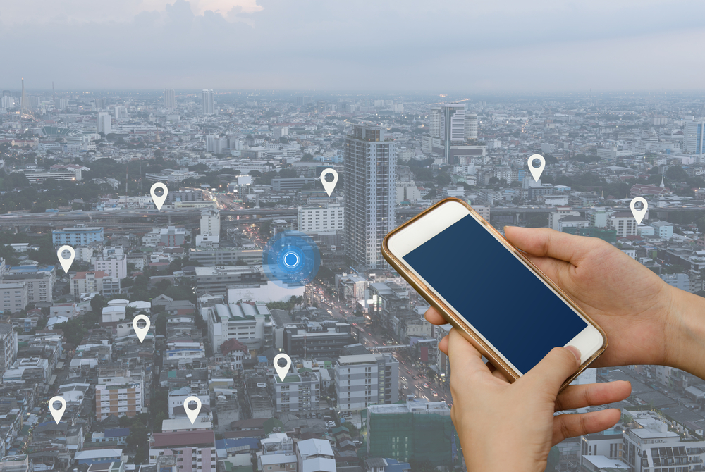 Shutterstock_529436278  Woman hand using mobile smartphone searching location on map and pin above blue tone city scape and network connection, internet of things, satellite navigation system app, smart journey concept