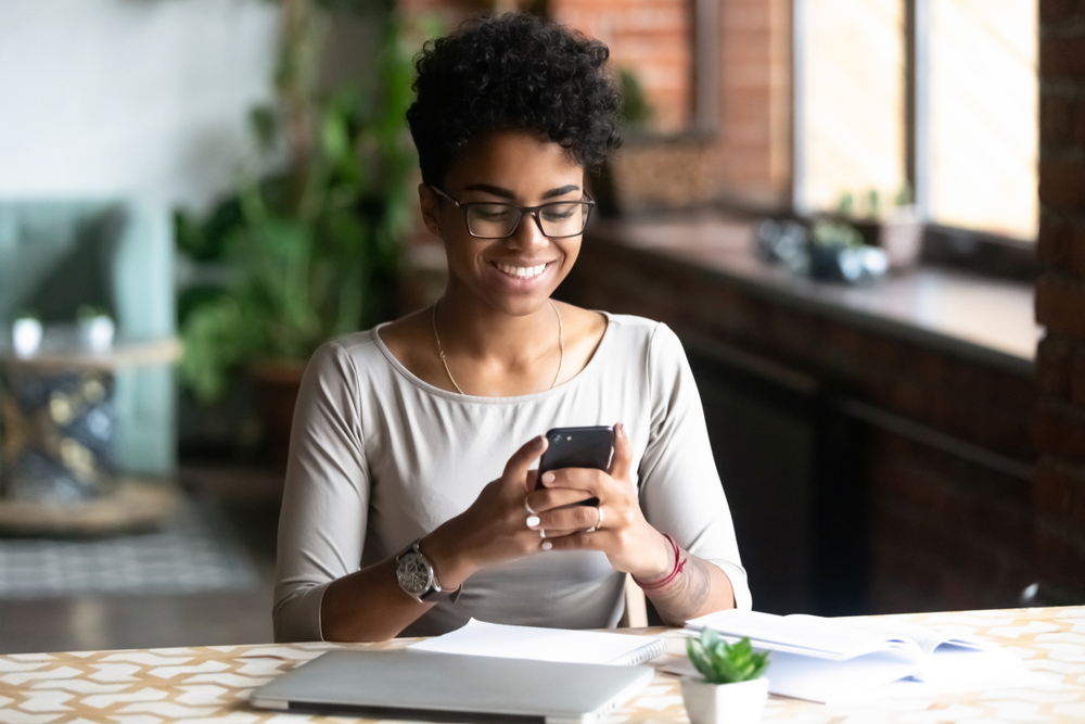 Shutterstock_1282956880 Cheerful african student black woman sitting at table studying using laptop reading a book, take a break holding mobile phone surfing internet received message from friend chatting about weekend plans