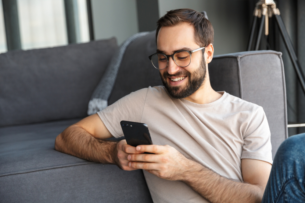 Shutterstock_1674151537 Attractive smart young man sitting on a floor in the living room, using mobile phone