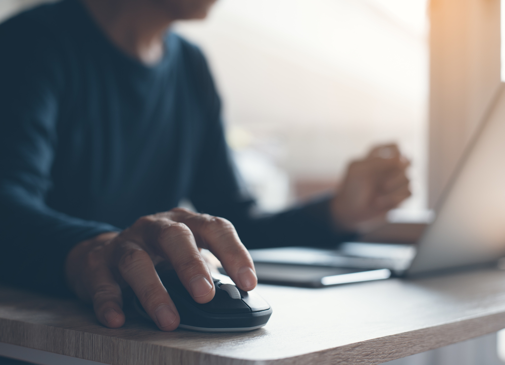 Shutterstock_631810814 Casual man, freelance working on laptop computer and clicking wireless digital mouse with digital tablet on office table, close up, dark tone