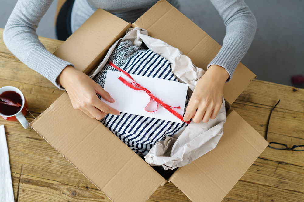 Shutterstock_510763483 Woman packaging a Christmas gift for posting tying a red ribbon around an envelope on a giftwrapped box in a cardboard carton, high angle view