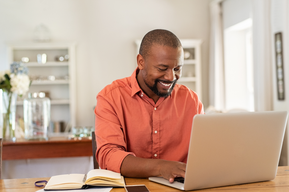 Shutterstock_1426545566 Smiling black man using laptop at home in living room. Happy mature businessman send email and working at home. African american freelancer typing on computer with paperworks and documents on table.