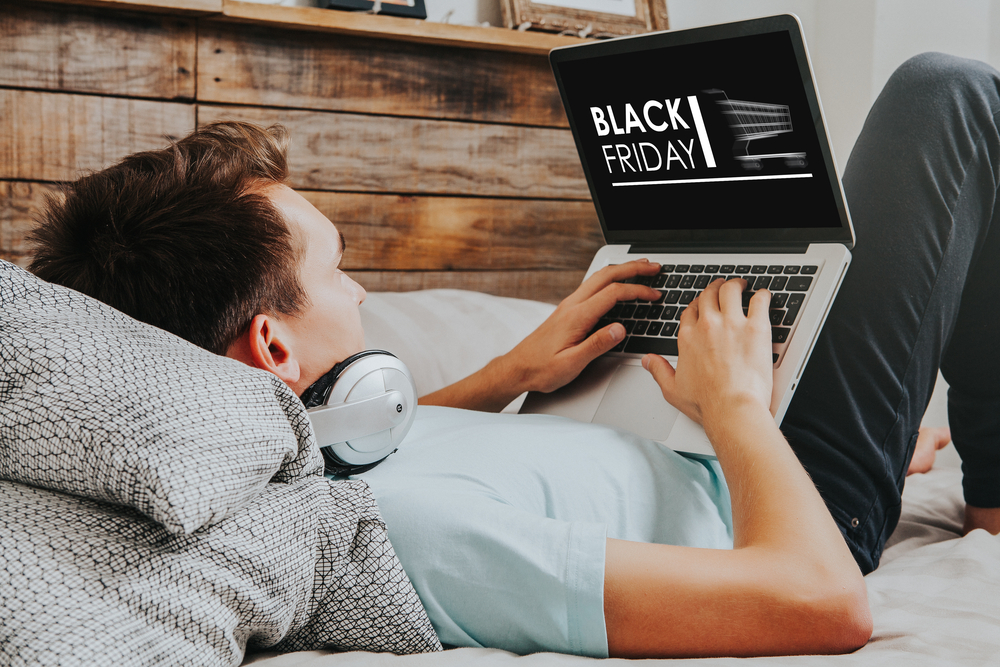 Shutterstock_747187003 Black Friday banner in a laptop computer while man uses it to buy by internet lying down at home.