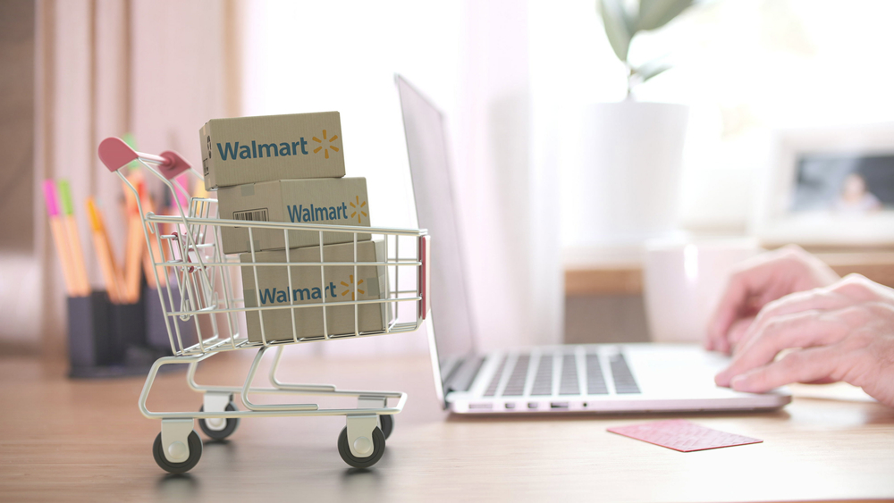 Shutterstock_1696873096 WALMART logo on boxes in shopping cart near the laptop. Editorial online shopping from home 3D rendering