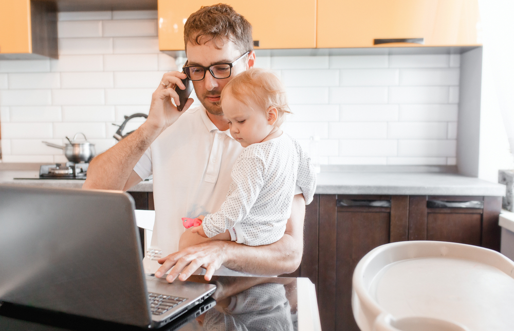 Shutterstock_1683011629 Handsome young man working at home with a laptop with a baby on his hands. Stay home concept. Home office with kids.