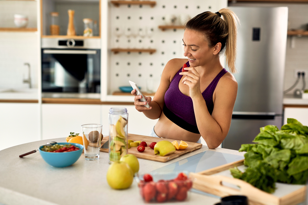 Shutterstock_1745003447 Young happy athletic woman eating fruit while text messaging on mobile phone in the kitchen.