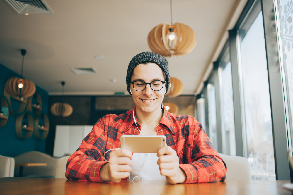 Shutterstock_562571530 Attractive young man is sitting in office and drinking tea. He is holding a mobile phone and watching video or reading interesting article. Handsome guy is smiling