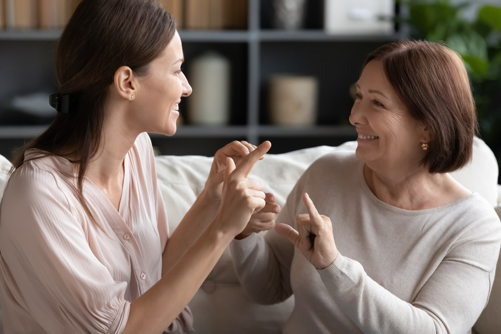 Shutterstock_1667439799 Head shot pleasant young dumb woman communicating nonverbal with deaf middle aged lady, sitting together on couch. Smiling impaired disabled two generations family using sign language at home.