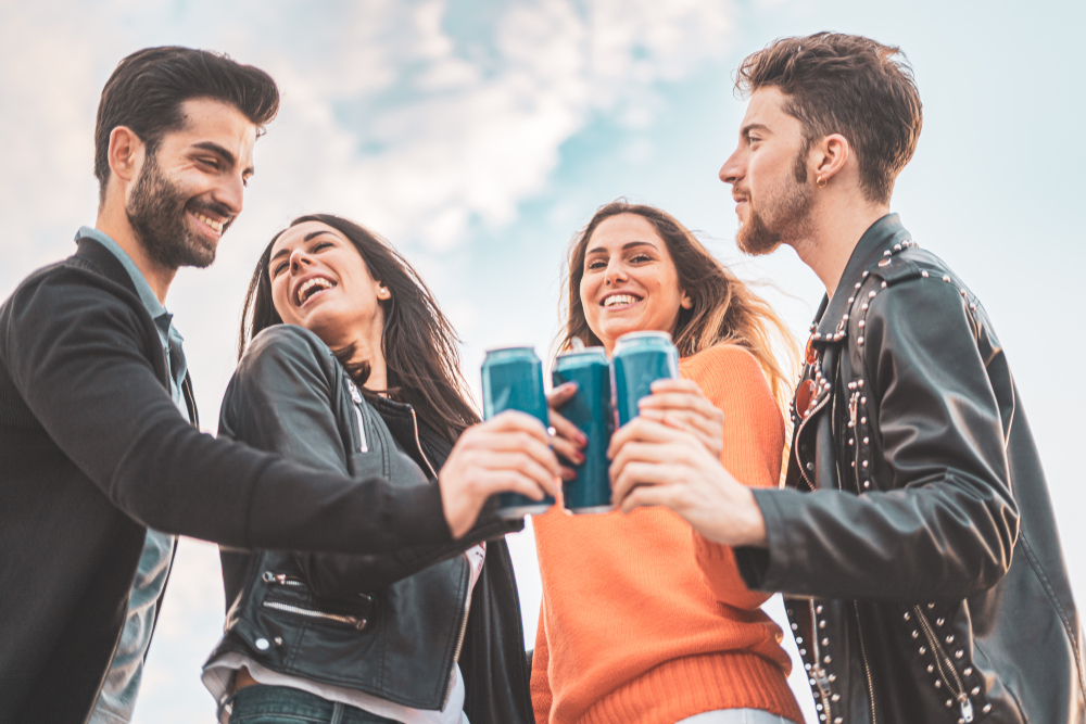 Shutterstock_1415669876 Two guys and two girls smiling and toasting with canned beers outdoor. Carefree youth having fun outdoors.