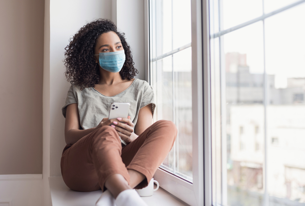 Shutterstock_1864147915 Sad woman alone during coronavirus pandemic wearing face mask indoors at home for social distancing. Mixed race girl looking at window. Anxiety, stress, lockdown, mental health crisis concept