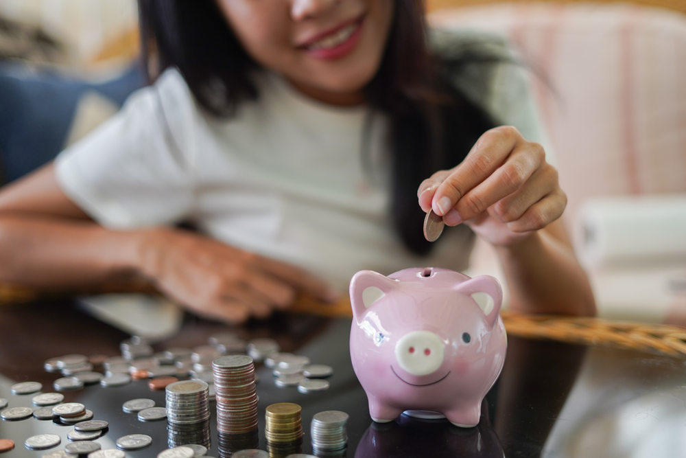 Shutterstock_1689229639 Woman is smiling and hand putting coin into piggy bank, Finance or Savings concept.