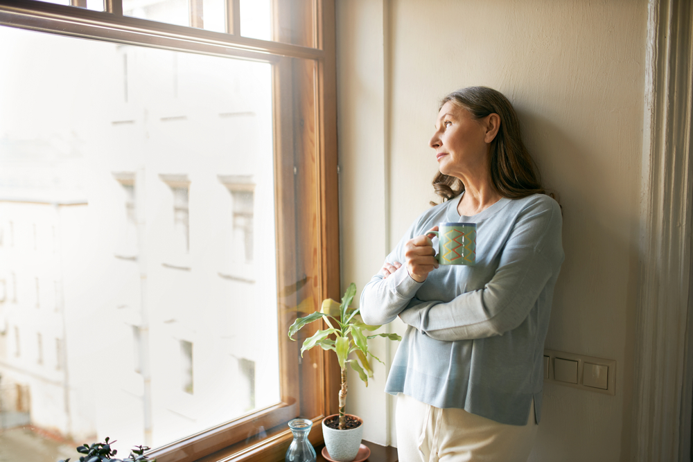 Shutterstock_1756606511 Portrait of thoughtful retired mature woman in casual clothes standing by large window, leaning on white wall, looking outside, drinking tea. People, social distancing and quarantine concept